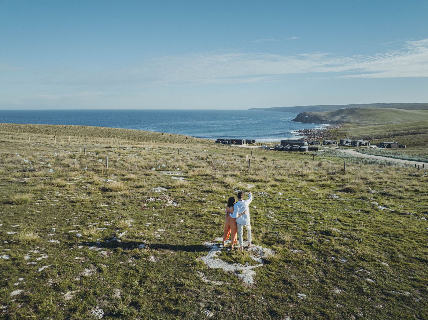 Two guests standing on a grassy coastal rise, looking down at a row of AUTRA retreats above the Southern Ocean